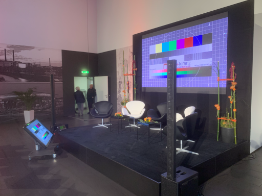 Leerer Bühnenbereich mit Stühlen und einem Bildschirm im Hintergrund.