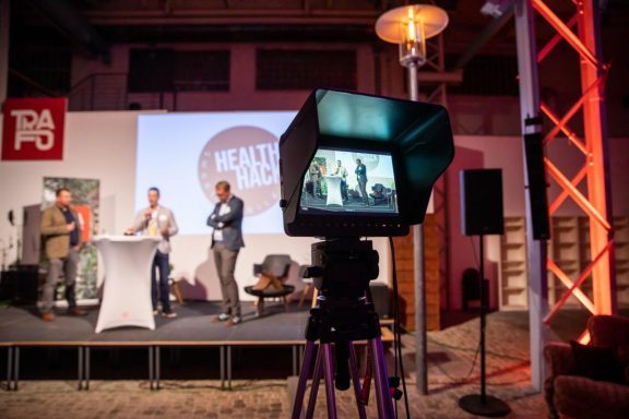 Auf einer Bühne präsentieren drei Personen vor einem Kameraviewer. Hintergrund leicht unscharf.